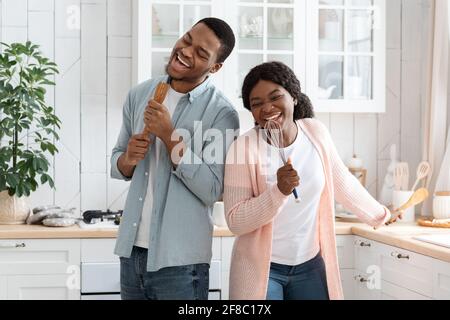 Inländisches Band. Fröhliche Afrikanische Ehepartner, Die In Der Küche Herumtollten Und Utensilien Als Mikrofone Verwendeten Stockfoto