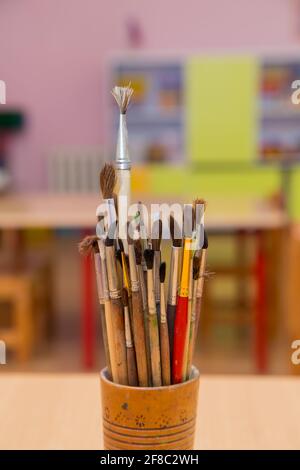 Künstlerische Pinsel stehen in einer Nahaufnahme aus Holzglas. Die Gegenstände für das Zeichnen mit den Farben werden zusammen gesammelt. Im Hintergrund sind Tisch und Schrank außer Fokus. Warmes, weiches Tageslicht. Stockfoto