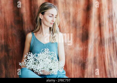 Verträumt attraktive weibliche Blondine in einem blauen Frühlingsjumpsuit mit einem Bouquet von Gypsophila-Blüten. Porträt einer selbstbewussten Frau in den Dreißigern. Sommer Stockfoto