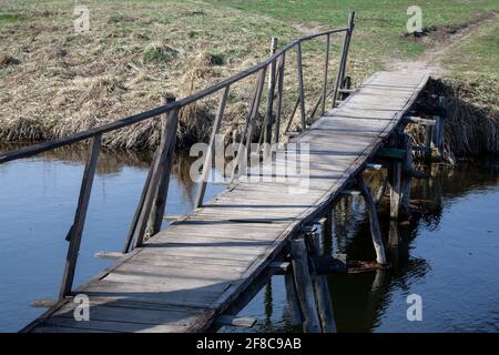 Alte Holzbrücke über einen Fluss. Verfallene Brücke. Zerstörte Stützelemente. Enger Fluss in einem Feld. Sonniger Tag. Schrecklich Stockfoto
