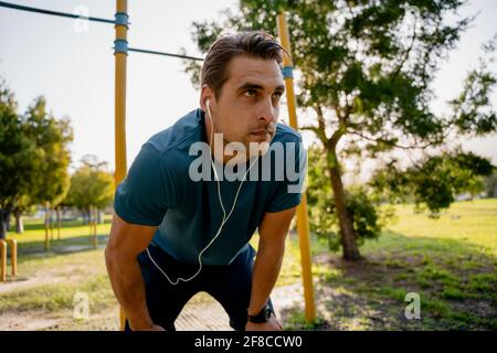 Fokussierter junger Athlet, der sich beugte, um sich zu strecken und zu hören Musik in Ohrhörern mit Smartwatch beim Training im Park Stockfoto