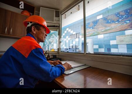 Kontrolleur in Wasserkraftwerk Maschinenraum.Senior Mann Arbeiter in roten Hardhat mit Papier Logbuch. Schaut durch das Fenster. Stockfoto