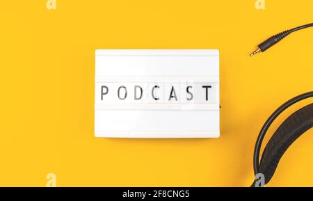 Podcast-Wort auf Leuchtkasten, gelber Tischhintergrund mit Kopfhörern, Draufsicht Foto Stockfoto