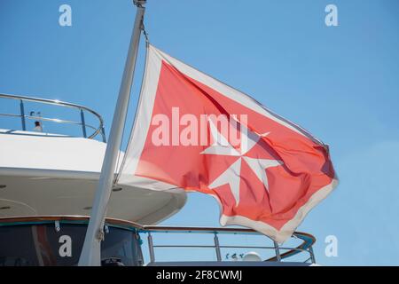 Flagge Maltas auf einem Luxusboot, das in der Marina in Griechenland vor Anker liegt. Maltesische alte Flagge, weißes Kreuz auf rotem Hintergrund, winkt am Heck der Yacht. Blauer Himmel Hintergrund, Stockfoto