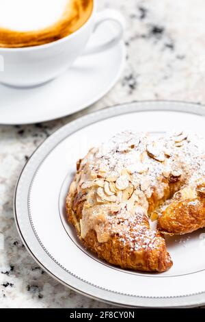 Mandelcroissant auf dem weißen Teller serviert zusammen mit einer Tasse Cappuccino-Kaffee für ein Frühstück oder eine Kaffeepause. Stockfoto