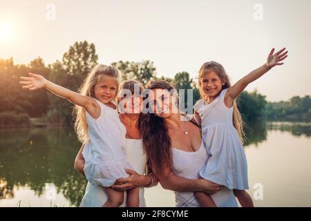 Muttertag. Familie mit Kindern, die am Sommerfluss spazieren gehen. Mutter und Großmutter hielten Mädchen fest und lachten, die Arme hochhebend. Drei Dgenerationen Stockfoto