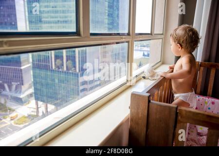 Ein charmantes Kleinkind blickt aus dem Fenster auf die Metropole, während es in einer Krippe steht. Baby in einer Krippe blickt aus dem Fenster auf die Stadt