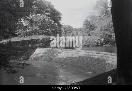 An einem Sommertag, historisch, Blick auf eine gestufte Wand oder ein Wehrwerk über einen Fluss. Diese von Menschen geschabekommen Struktur ist eine Barriere über die Breite eines Flusses, die sowohl die Höhe des Flusses Ebene und den Fluss des Wassers ändert. Sie werden oft gebaut, um Überschwemmungen zu verhindern. Staudämme und Stauwehre werden häufig im gleichen Kontext eingesetzt, unterscheiden sich jedoch von Staudämmen dadurch, dass sie in erster Linie so ausgelegt sind, dass das Wasser über das Wehrwerk, also die Barriere, fließt und nicht hinter einer Wand zurückgehalten wird. Stockfoto