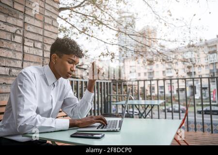 Ein schwarzer Mann sitzt auf einer Sommerterrasse oder Veranda in der Nähe eines Cafés. Der Typ arbeitet an einem Laptop. Blick auf das Frühlingsgeschäft in der Innenstadt Stockfoto