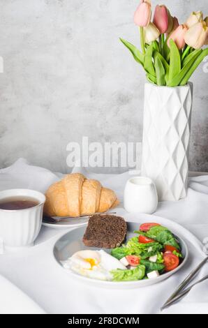 Frühmorgendliche Tischdekoration. Salat auf dem Teller, Ei, Tasse Kaffee und Croissant, frische Tulpen in Vase auf weißem Tischtuch. Stockfoto