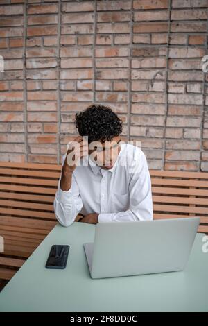 Vertikales Foto EIN schwarzer Mann sitzt auf einer Sommerterrasse oder Veranda in der Nähe eines Cafés. Der Typ arbeitet an einem Laptop und nutzt ein Smartphone. Blick auf den Frühling Stockfoto
