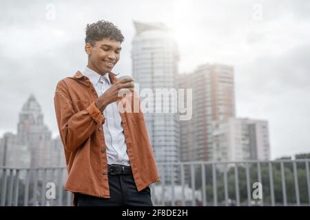 Ein junger schwarzer Mann in einer braunen Jacke hält einen Drink in einer Tasse in seinen Händen vor dem Hintergrund einer städtischen Skyline oder der Innenstadt. Der Typ trinkt Stockfoto
