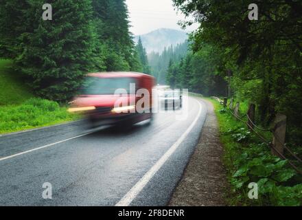 Verschwommene Autos auf der Straße im nebligen Wald an regnerischen Tagen im Sommer. Wunderschöne Bergstraße, Bäume, grünes Laub im Nebel. Landschaft mit rotem Auto in Mo Stockfoto