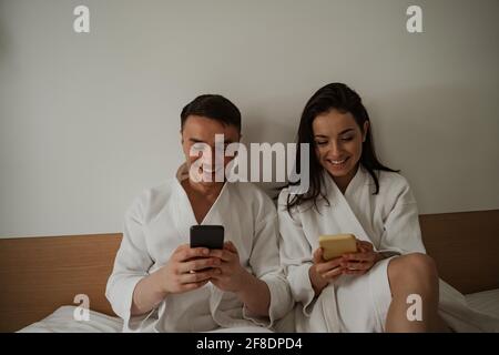 Morgen des glücklichen jungen Paares in weißen Bademänteln im Bett zu Hause, mit dem Telefon Stockfoto