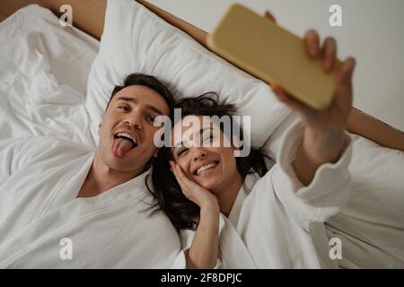 Morgen von glücklichen jungen Paar in weißen Bademänteln im Bett zu Hause, machen Selfie Stockfoto