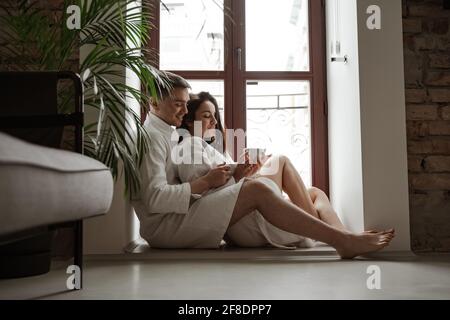 Morgen mit einem glücklichen jungen Paar in weißen Bademänteln zu Hause, das am Fenster sitzt, Kaffee trinkt und umarmt Stockfoto