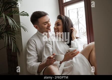 Morgen mit einem glücklichen jungen Paar in weißen Bademänteln zu Hause, das am Fenster sitzt, Kaffee trinkt und umarmt Stockfoto