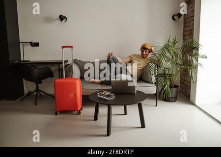 Der junge Mann, der unterwegs ist, sitzt auf der Couch und schaut sich in den Sommerferien etwas auf dem Laptop im Hotelzimmer an. Cooler Mann in lässiger Kleidung mit Gepäck Stockfoto