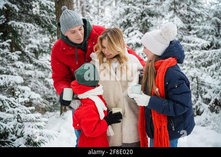 Vater, Mutter, Sohn und Tochter lachen und gehen gemeinsam im verschneiten Wald. Sie trinken Tee aus einer Thermoskanne und Tassen, um sich warm zu halten Stockfoto