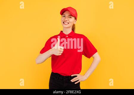 Fröhliche junge Kurierin, die eine rote Uniform trägt und ihren Daumen nach oben zeigt, vor einem gelben Hintergrund stehend, Lieferservice Stockfoto