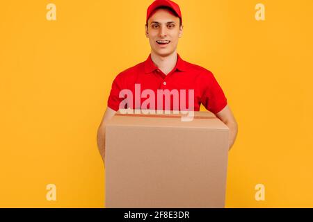 Fröhlicher, aufgeregt männlicher Kurier mit rotem T-Shirt und Mütze, der das Paket auf einem isolierten gelben Hintergrund hält, Express-Lieferung, Einkaufskonzept Stockfoto