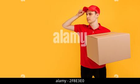 Fröhlicher, aufgeregt männlicher Kurier mit rotem T-Shirt und Mütze, der das Paket auf einem isolierten gelben Hintergrund hält, Express-Lieferung, Einkaufskonzept Stockfoto