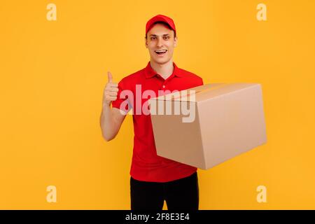 Fröhlicher, aufgeregt männlicher Kurier mit rotem T-Shirt und Mütze, der das Paket auf einem isolierten gelben Hintergrund hält, Express-Lieferung, Einkaufskonzept Stockfoto