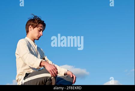 Kaukasischer Mann aus Spanien in einem beigefarbenen Hemd meditiert in Eine sitzende Position auf bewölktem Himmel Hintergrund Stockfoto