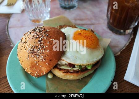Frühstückssandwich mit einem Ei auf der Sonnenseite in einem Brioche-Brötchen mit allem Gewürz und Mohnsamen und auf einen blaugrün gefärbten Teller gelegt. Brunch Stockfoto