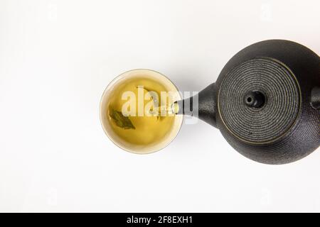 Heißer grüner Tee, der von einer Teekannen zu einer keramischen Teetasse gegossen wird, isoliert auf weißem Hintergrund Stockfoto