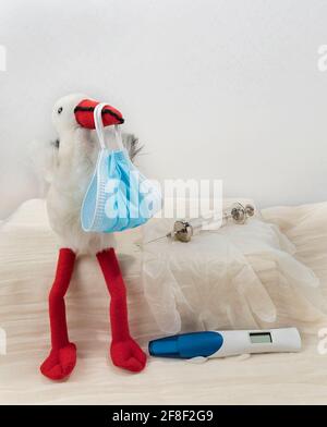 Konzeptuelles Bild, wie Covid-19 Schwangere und den Muttermund beeinflusst. Storchenspielzeug mit Bundle aus Gesichtsmaske, Latexhandschuhen, Spritze mit Nadel und Schwangerschaft Stockfoto