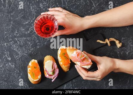 Weibliche Hände halten Bruschetta-Sortiment mit Baguette, Frischkäse, rotem Kaviar, Lachs und einem Glas Rotwein auf dem Steinhintergrund. Set von Appetiz Stockfoto