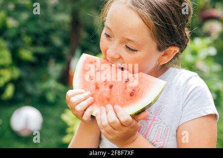 Porträt eines hübschen Mädchens, 7-8 Jahre alt, das im Garten ein Stück Wassermelone auf dem Gras frisst. Sommerlicher Lifestyle Stockfoto