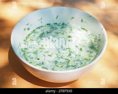 Weiße Platte mit Okroshka steht auf Holzoberfläche im Freien in Sonnenlicht Stockfoto