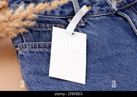 Makro-Modell mit weißem, leerem Papieretikett oder Etikett Eine Nadel für das Logo auf einer blauen Jeans mit einem Einrichtung aus trockenem Pampa-Gras oder Schilf Stockfoto