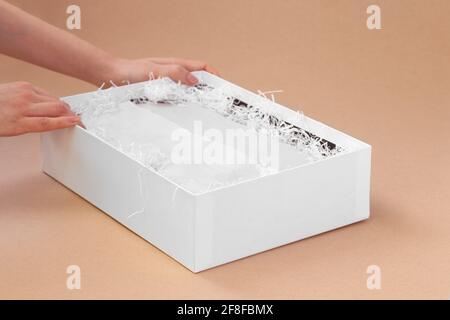 Die Hände einer Frau packen für einen Kunden eine weiße Schachtel Mit Kleidung aus weißem Seidenpapier und geschreddertem Papier auf Ein beigefarbener Papierhintergrund Stockfoto