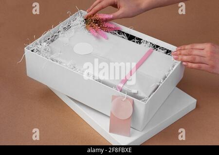 Die Hände einer Frau packen eine Schachtel mit Kleidung und einem aus Rosafarbenes, leeres Etikett für eine Marke oder ein Logo und ein Einrichtung mit rosa Blumen auf beigefarbenem Papierhintergrund Stockfoto