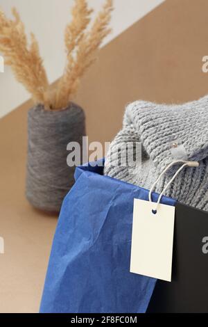 Makroweißes leeres Papier Logo-Tag auf einem grauen gestrickten Schal in einer schwarzen Box und getrocknete Pampas Gras in Eine Vase aus Spulengarn auf beigefarbenem Papier Stockfoto