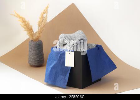 Weißes, leeres Papier-Logo auf einem grauen Strickschal In einer schwarzen Schachtel und blauem Seidenpapier und getrocknet pampas Gras in einer Vase aus Spulengarn auf beigefarbenem Papier Stockfoto