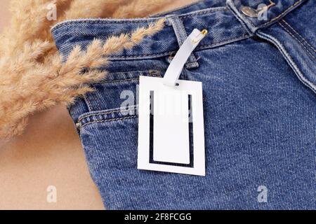 Aufgedrucktes weißes, leeres Papieretikett oder Etikett mit geometrischem Muster Logoschlitze auf blauer Jeans mit trockenem Pampagras Einrichtung Stockfoto