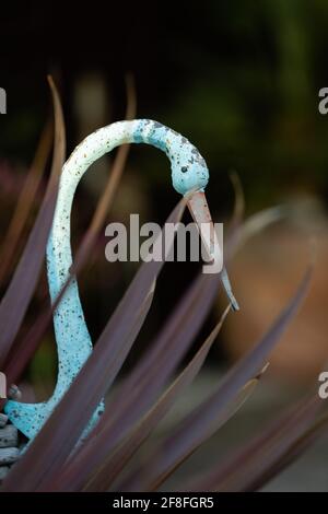 Blau bemalter Zierreiher mit Holzfedern im Garten mit Fächerförmige cordyline australis „Red Star“ Stockfoto
