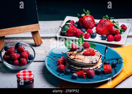 Köstliches Dessert, Pfannkuchen mit dunklem Schokoladensirup und frisches Obst mit Minze auf blauem Teller und Holztisch. Schwarzer Hintergrund für Kopierraum Stockfoto