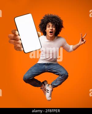 Aufgeregt afroamerikanischen Teenager Kerl springen mit Smartphone, Demonstration leeren Bildschirm auf orangenen Hintergrund Stockfoto