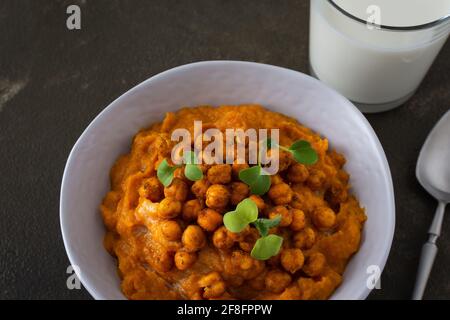 Dicke Kürbissuppe mit gebackenen Kichererbsen und Rettichsprossen auf einem Teller mit einem Glas Milch auf dem Tisch, leckeres gesundes Abendessen Stockfoto