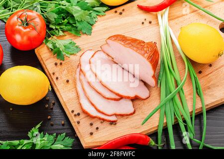 Ein Blick von oben auf dünn geschnittenes ​​smoked Schweinefleisch auf einem Holzbrett neben Petersilie, grünen Zwiebeln, schwarzen Erbsen, Tomaten, Zitronen und Paprika. Stockfoto