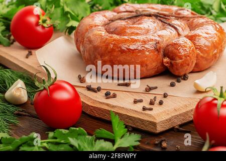 Seitenansicht von geräucherter hausgemachter Wurst, gebunden mit einer Peitschenspirale auf einem Holzschneidebrett. Tomaten, Knoblauch, Petersilie, Paprika, Nelken sind in der Nähe. Schließen-U Stockfoto