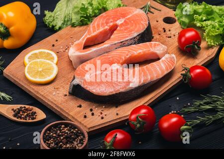 Zwei saftige, appetitliche Lachssteaks werden auf einem Holzbrett neben Gewürzen, Rosmarin, Zitrone, Tomaten, Salat und Meersalz ausgelegt. Nahaufnahme, oben und s Stockfoto