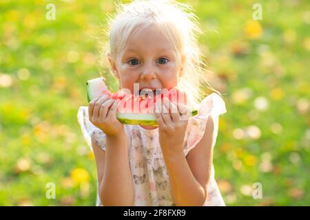 Cute blonde kleine Mädchen essen Wassermelone auf dem Gras im Park. Stockfoto
