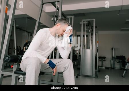 Sehr müde nach einem intensiven Training sitzt ein Mann im Fitnessstudio und wischt sich mit einem Handtuch Schweiß vom Gesicht. Stockfoto