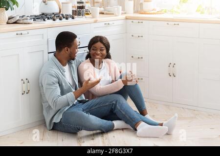 Glücklicher Schwarzer Mann Und Frau Sitzen Auf Dem Boden In Der Küche Und Chatten Stockfoto